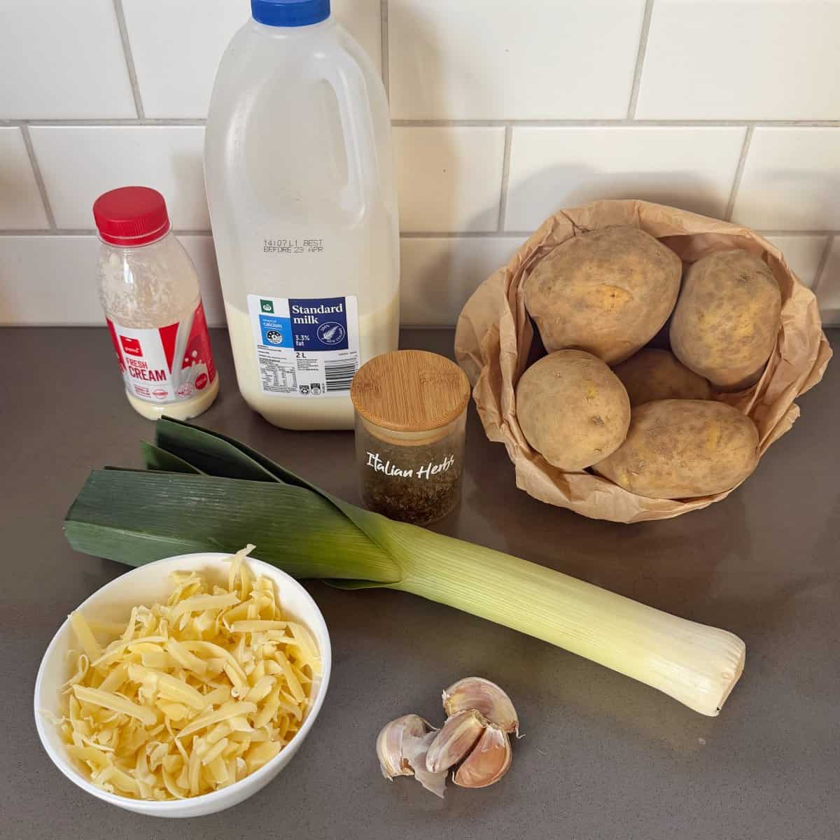 Ingredients on a counter: milk, double cream, potatoes, Italian herbs, leeks, shredded cheese, and garlic, arranged in front of a white tiled backsplash.