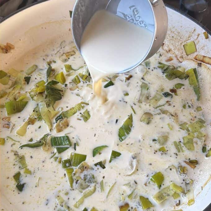 Potato Leek Gratin Milk is being poured from a measuring cup into a pan of simmering leeks and herbs.