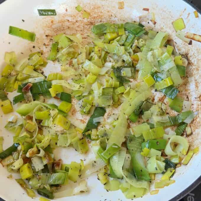 Potato Leek Gratin Chopped leeks sautéing in oil and herbs in a white frying pan, with browning visible on the pan and some pieces.