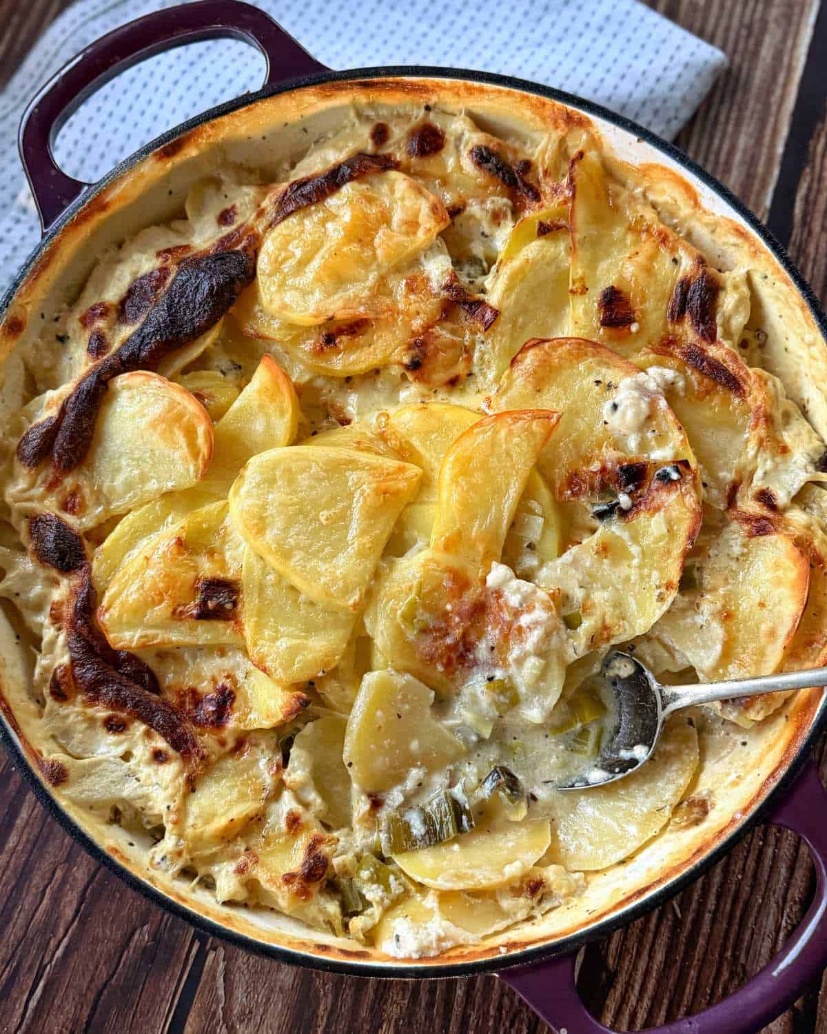A round casserole dish filled with creamy, baked scalloped potatoes, with a serving spoon scooping out a portion. The dish rests on a wooden surface with a cloth nearby.