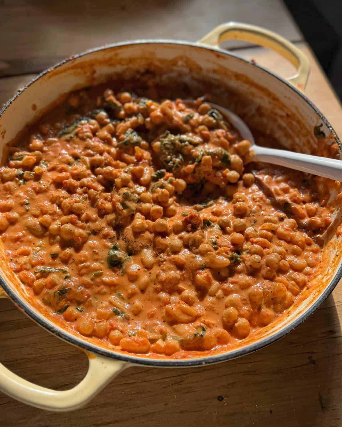 Marry Me Chickpeas and Beans A pot of creamy beans in a tomato-based sauce with herbs, shown on a wooden surface with a serving spoon inside.