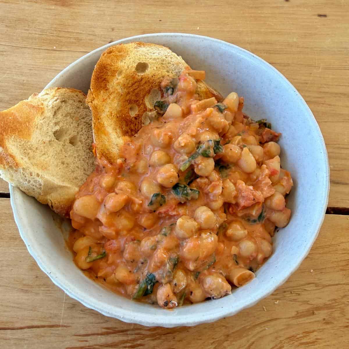 Marry Me Chickpeas and Beans A bowl of creamy white beans in tomato sauce with herbs, served with two slices of toasted bread on a wooden table.