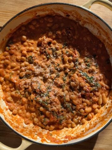 A round cream-colored pot filled with baked beans in a tomato-based sauce, garnished with herbs, sits on a wooden surface in natural sunlight.