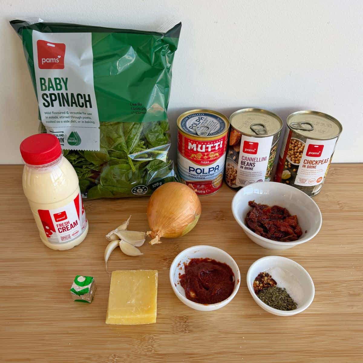 Marry Me Chickpeas and Beans Ingredients including baby spinach, canned tomatoes, beans, chickpeas, cream, onion, garlic, vegetable stock, cheese, tomato paste, dried herbs, spices, and sun-dried tomatoes on a wooden surface.