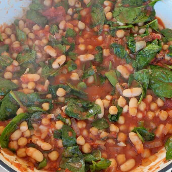 Marry Me Chickpeas and Beans White beans, spinach, and diced tomatoes simmer in a tomato-based sauce, creating a colorful and hearty vegetable stew in a pot.