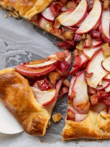 A rustic fruit tart with sliced apples and rhubarb sits on parchment paper, with one slice being lifted by a white pie server.