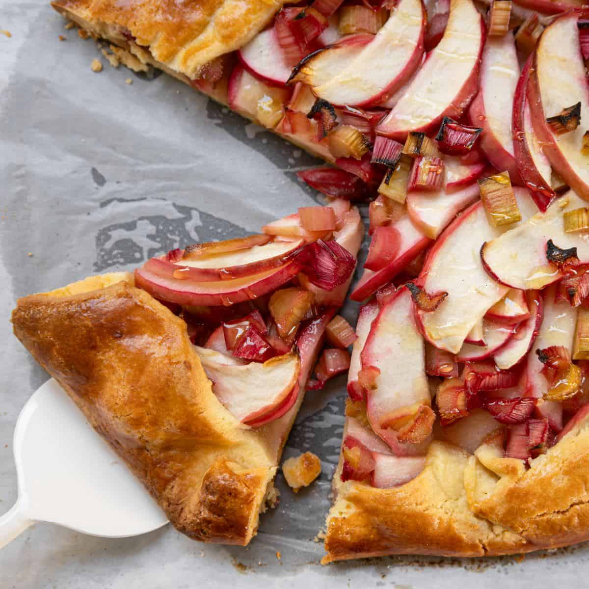 Rhubarb and Apple Galette A rustic fruit tart with sliced apples and rhubarb sits on parchment paper, with one slice being lifted by a white pie server.
