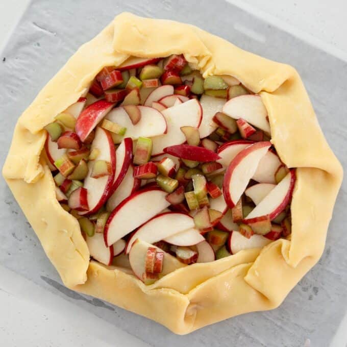 Rhubarb and Apple Galette Unbaked galette with sliced apples and chopped rhubarb on a lined baking sheet, edges of the pastry folded inward.