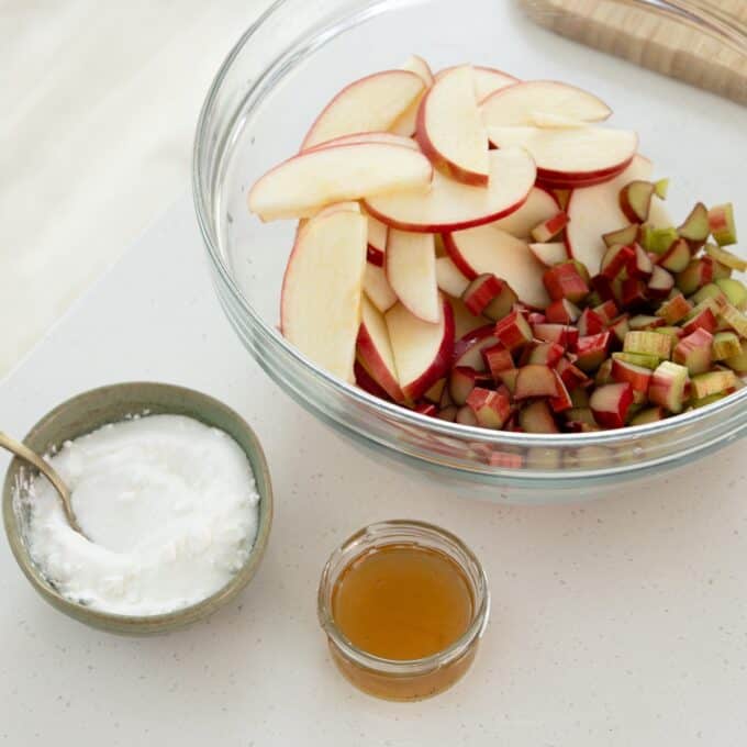 Rhubarb and Apple Galette A glass bowl of sliced apples and diced rhubarb beside a small bowl of sugar and a jar of honey on a white countertop.