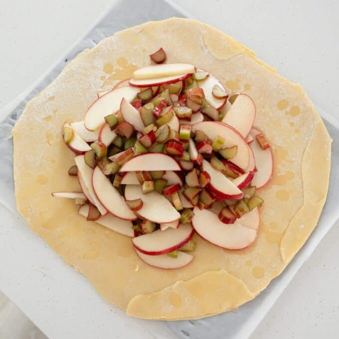 Rhubarb and Apple Galette Unbaked galette with sliced apples and chopped rhubarb on rolled dough, topped with a light drizzle, placed on parchment paper.