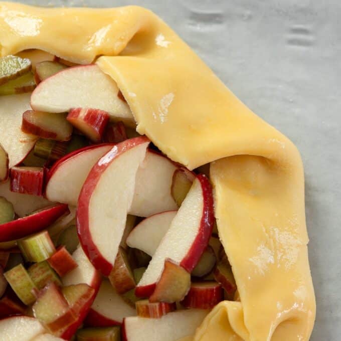 Rhubarb and Apple Galette Unbaked galette with sliced red apples and rhubarb pieces on a crust, edges folded over, on a baking sheet.
