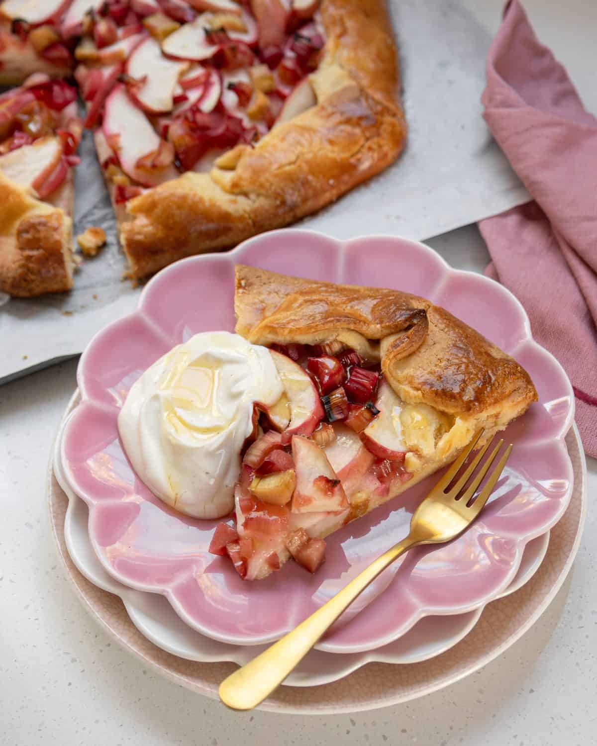 Rhubarb and Apple Galette A slice of fruit galette with whipped cream and honey is served on a pink plate with a gold fork; the rest of the galette is in the background.