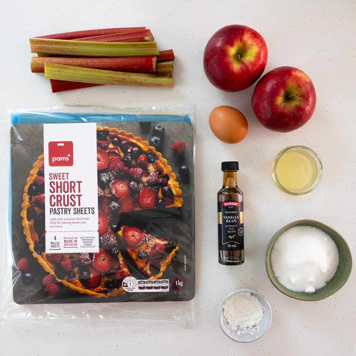 Rhubarb and Apple Galette Pastry sheets, rhubarb stalks, two apples, two eggs, vanilla extract, sugar, lemon juice, and a small bowl of flour on a countertop.