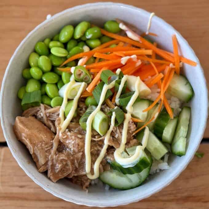 Slow Cooker Asian Style Chicken A rice bowl topped with shredded chicken, edamame, cucumber, shredded carrot, sliced green onions, and mayonnaise drizzle, served in a white bowl on a wooden surface.