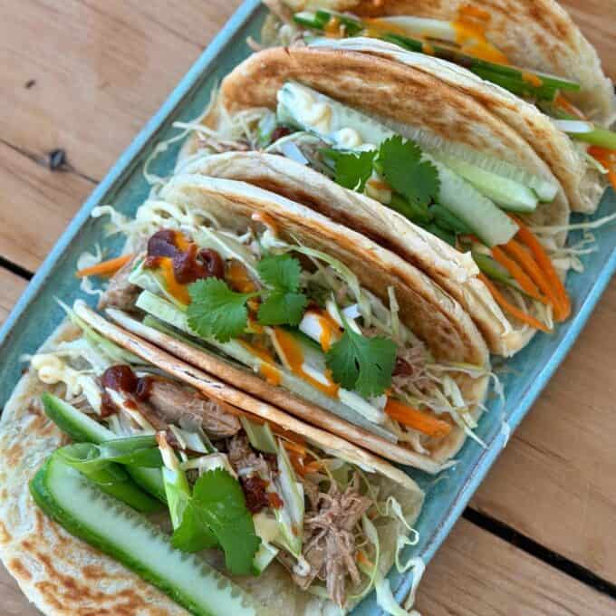 Slow Cooker Asian Style Chicken Four tacos filled with shredded meat, sliced cucumber, carrots, cabbage, cilantro, and sauces are arranged on a rectangular blue plate on a wooden table.