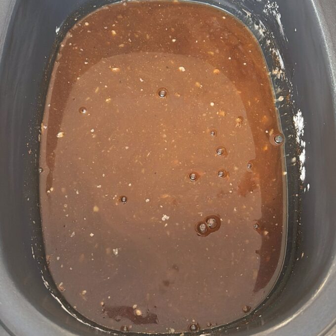Slow Cooker Asian Style Chicken A close-up of a dark brown liquid mixture with small bubbles and specks, contained in a black rectangular pan or slow cooker.