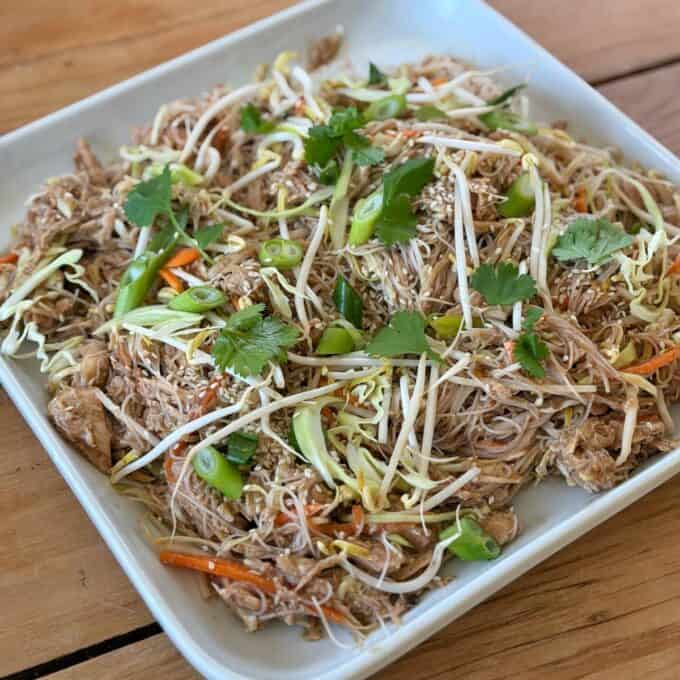 Slow Cooker Asian Style Chicken Plate of stir-fried noodles with shredded meat, bean sprouts, carrot strips, green onions, cilantro, and sesame seeds on a white rectangular dish.
