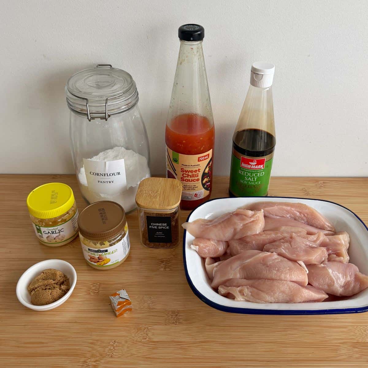 Slow Cooker Asian Style Chicken A tray of raw chicken breast fillets sits on a wooden surface beside jars and bottles of cornflour, sweet chili sauce, soy sauce, brown sugar, ginger, garlic, and a stock cube.