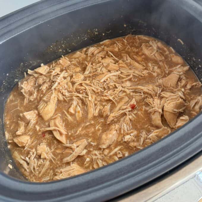 Slow Cooker Asian Style Chicken Shredded chicken in a brown sauce is cooking in a black slow cooker, with steam rising from the food.