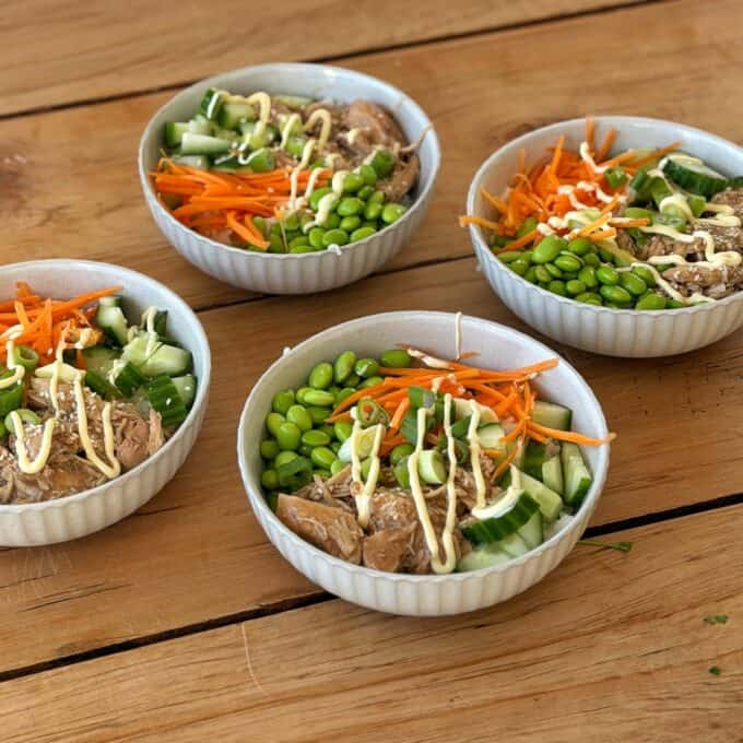 Slow Cooker Asian Style Chicken Four bowls of rice topped with shredded meat, edamame, cucumber, carrot, sesame seeds, and drizzled sauce, arranged on a wooden table.