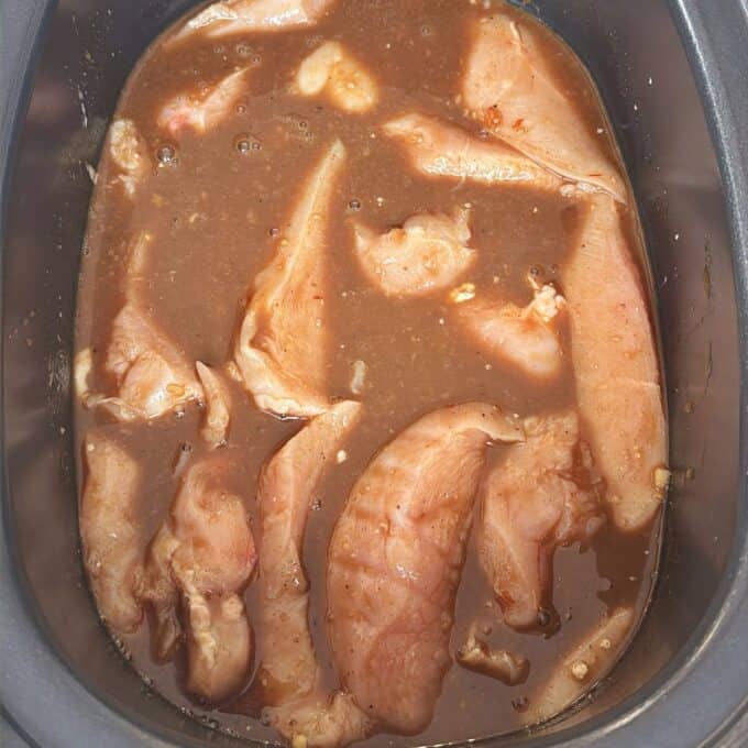 Slow Cooker Asian Style Chicken Raw chicken breast strips marinating in a brown liquid with visible seasonings, placed inside a slow cooker or similar container.