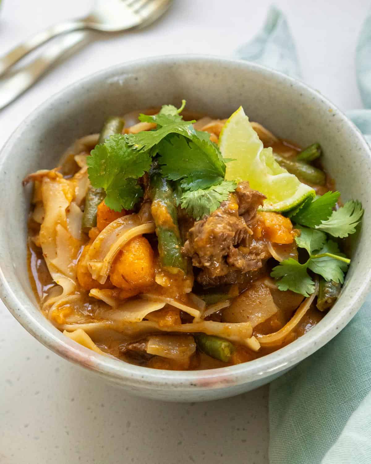 Slow Cooker Thai Beef Curry A bowl of noodle soup with beef, green beans, cilantro, lime, and vegetables, served in a gray bowl on a white surface.