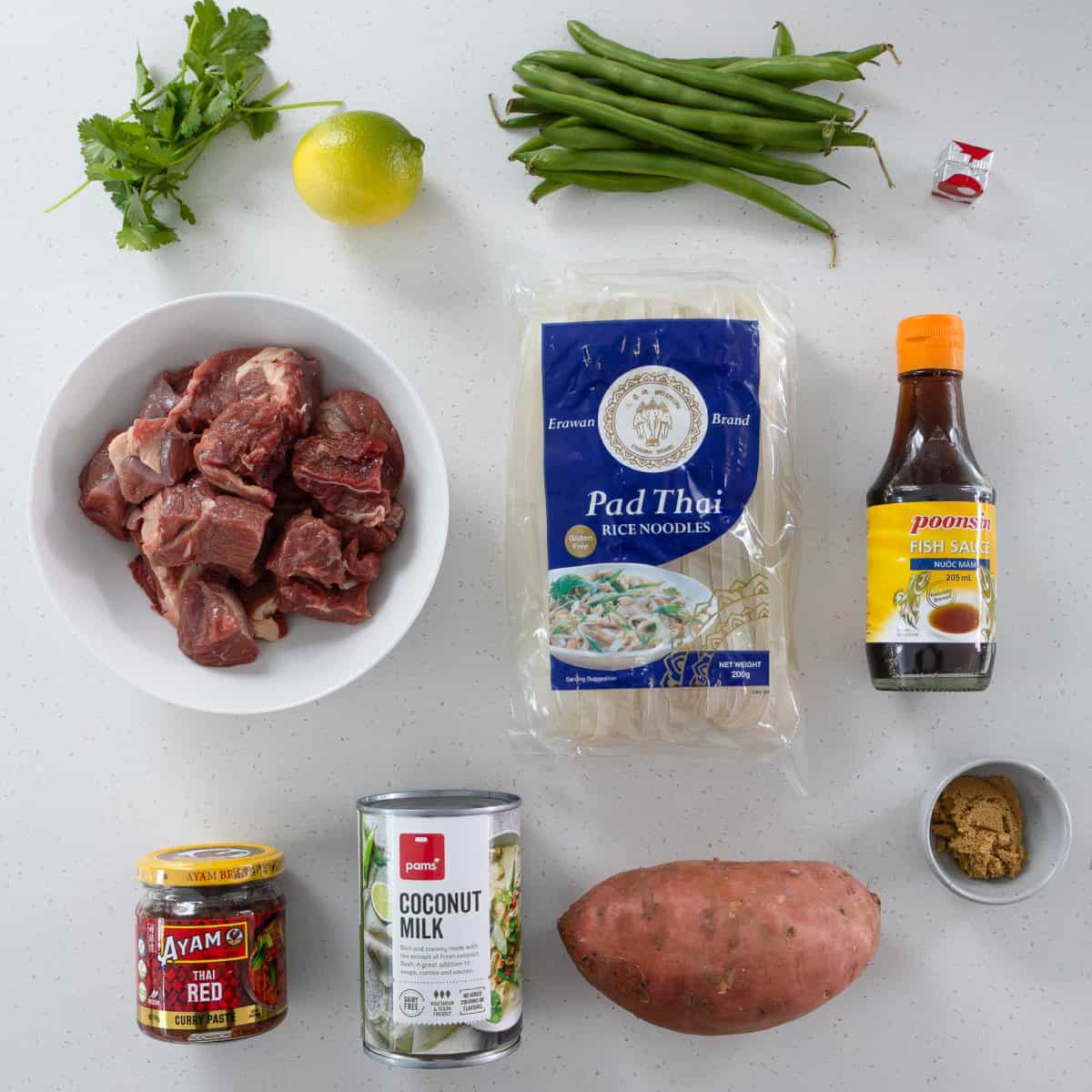 Slow Cooker Thai Beef Curry Various ingredients on a white surface, including raw beef, rice noodles, fish sauce, coconut milk, green beans, sweet potato, coriander, lime, curry paste, brown sugar, and a bouillon cube.