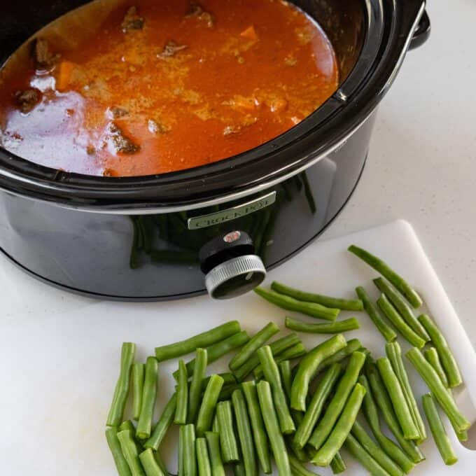 Slow Cooker Thai Beef Curry A slow cooker with red stew inside sits next to a cutting board topped with chopped fresh green beans.