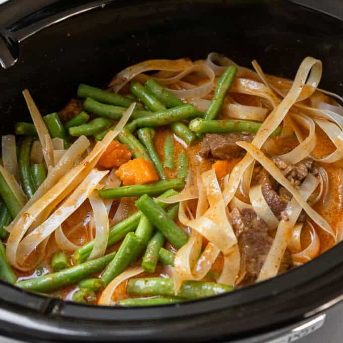 Slow Cooker Thai Beef Curry A slow cooker filled with stew containing flat noodles, green beans, chunks of meat, and carrots in a reddish broth.