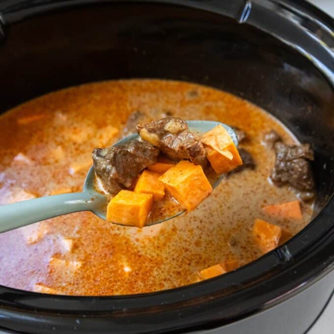 Slow Cooker Thai Beef Curry A spoon lifts chunks of beef and sweet potato from a creamy, orange-colored stew inside a slow cooker.