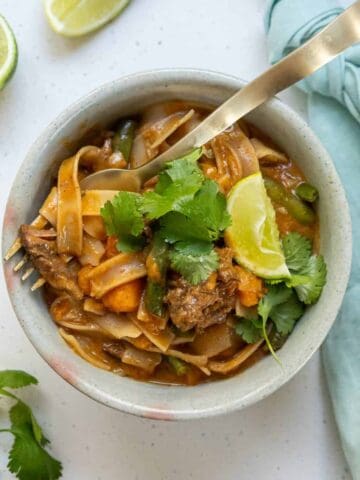 Bowl of noodle curry with beef, vegetables, fresh cilantro, and a lime wedge. A fork is placed in the bowl, and a blue napkin and lime slices are nearby.
