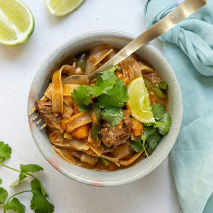 Slow Cooker Thai Beef Curry Bowl of noodle curry with beef, vegetables, fresh cilantro, and a lime wedge. A fork is placed in the bowl, and a blue napkin and lime slices are nearby.