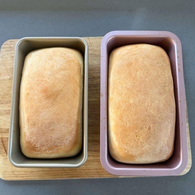 White Sandwich Bread Two loaves of baked bread sit in metal pans on a wooden cutting board, viewed from above.
