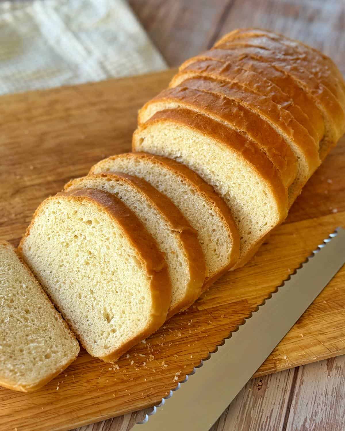 A loaf of white bread sliced on a wooden cutting board with a serrated bread knife beside it.