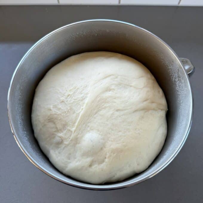 White Sandwich Bread A large ball of risen bread dough sits in a metal mixing bowl on a gray countertop.