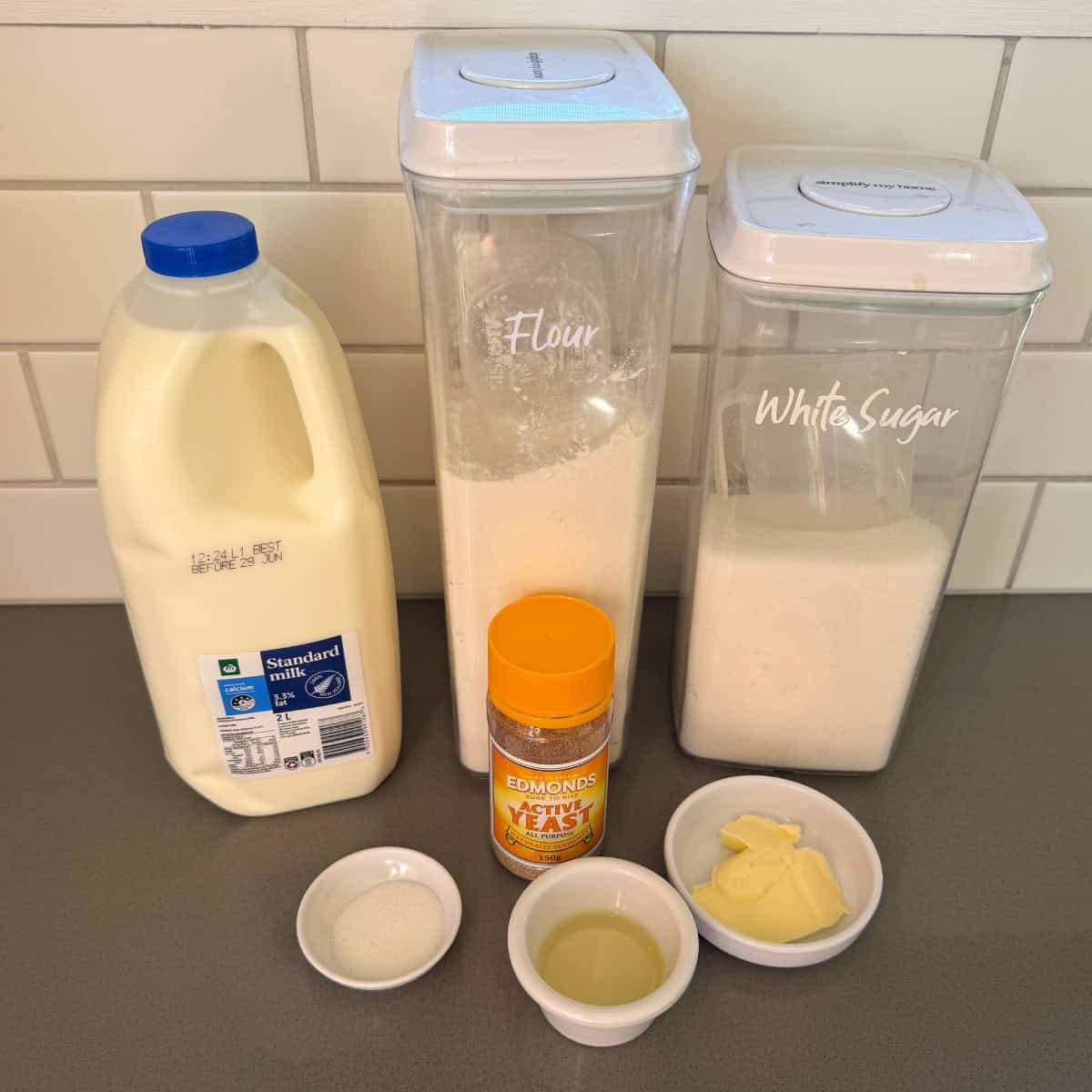 Baking ingredients on a counter: a bottle of milk, containers labeled flour and white sugar, yeast jar, small bowls with salt, oil, and butter. White tiled wall in the background.