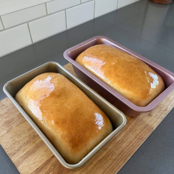 White Sandwich Bread Two loaves of shiny, golden brown bread sit in metal pans on a wooden cutting board, with a tiled backsplash in the background.