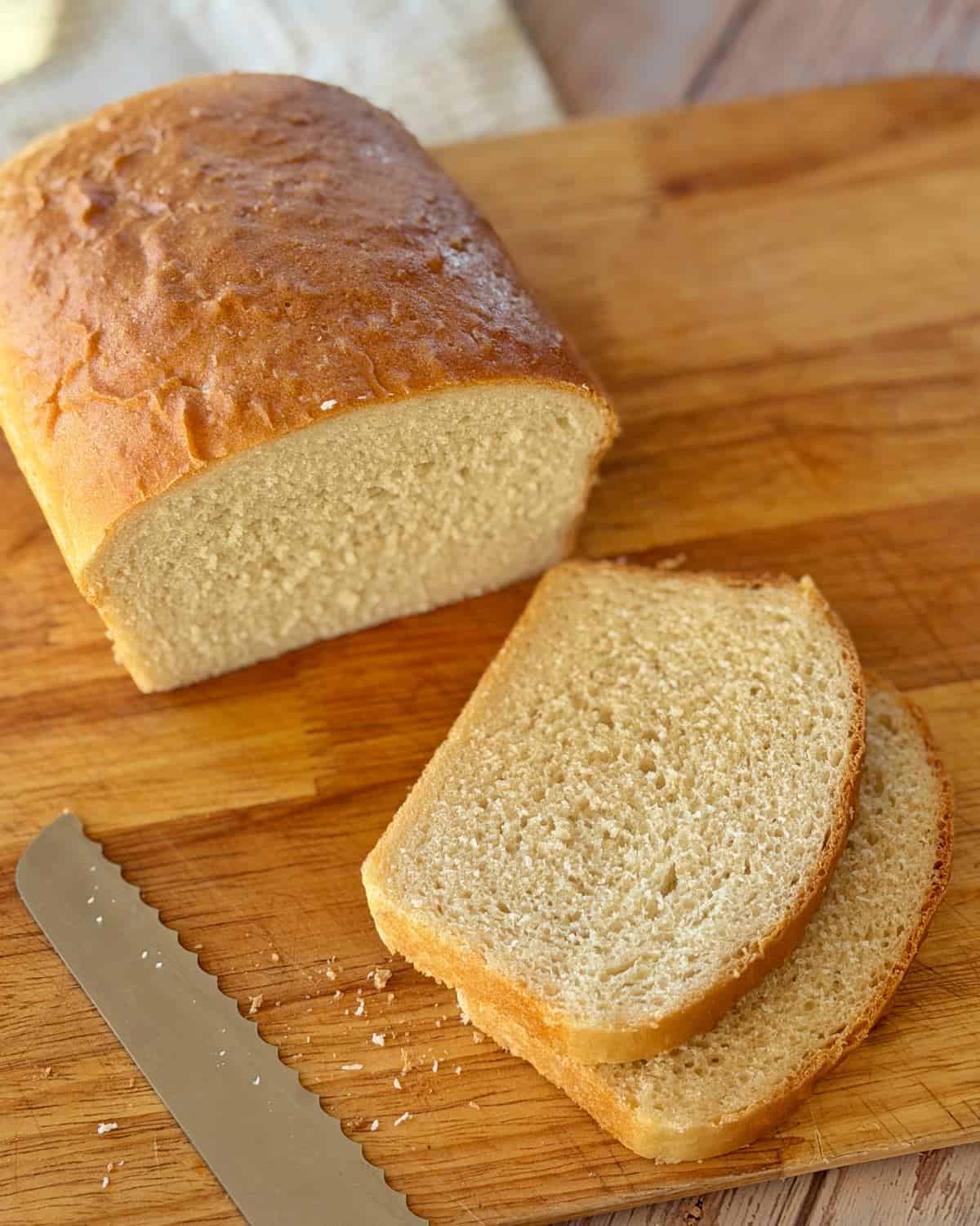 White Sandwich Bread A loaf of bread with two slices cut, resting on a wooden cutting board next to a serrated bread knife.