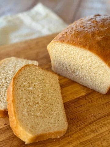 A loaf of white bread sits on a wooden cutting board with two slices cut and placed next to it.