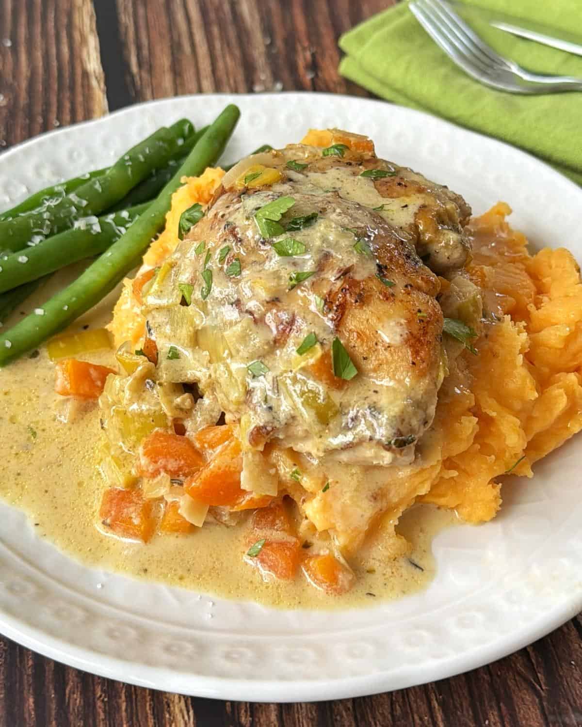 Creamy French Mustard Chicken Chicken with creamy vegetable sauce served over mashed sweet potatoes, accompanied by green beans on a white plate, with a fork and green napkin in the background.
