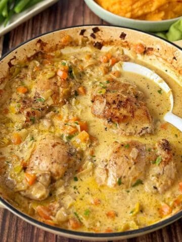 Creamy chicken thighs with carrots and onions cooked in a skillet, served with a white spoon. Mashed sweet potatoes and green beans are in the background.