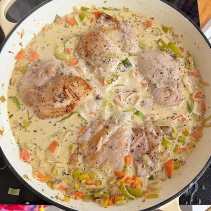 Creamy French Mustard Chicken Four seasoned chicken thighs simmer in a creamy sauce with chopped leeks and carrots in a white pan on a stovetop.