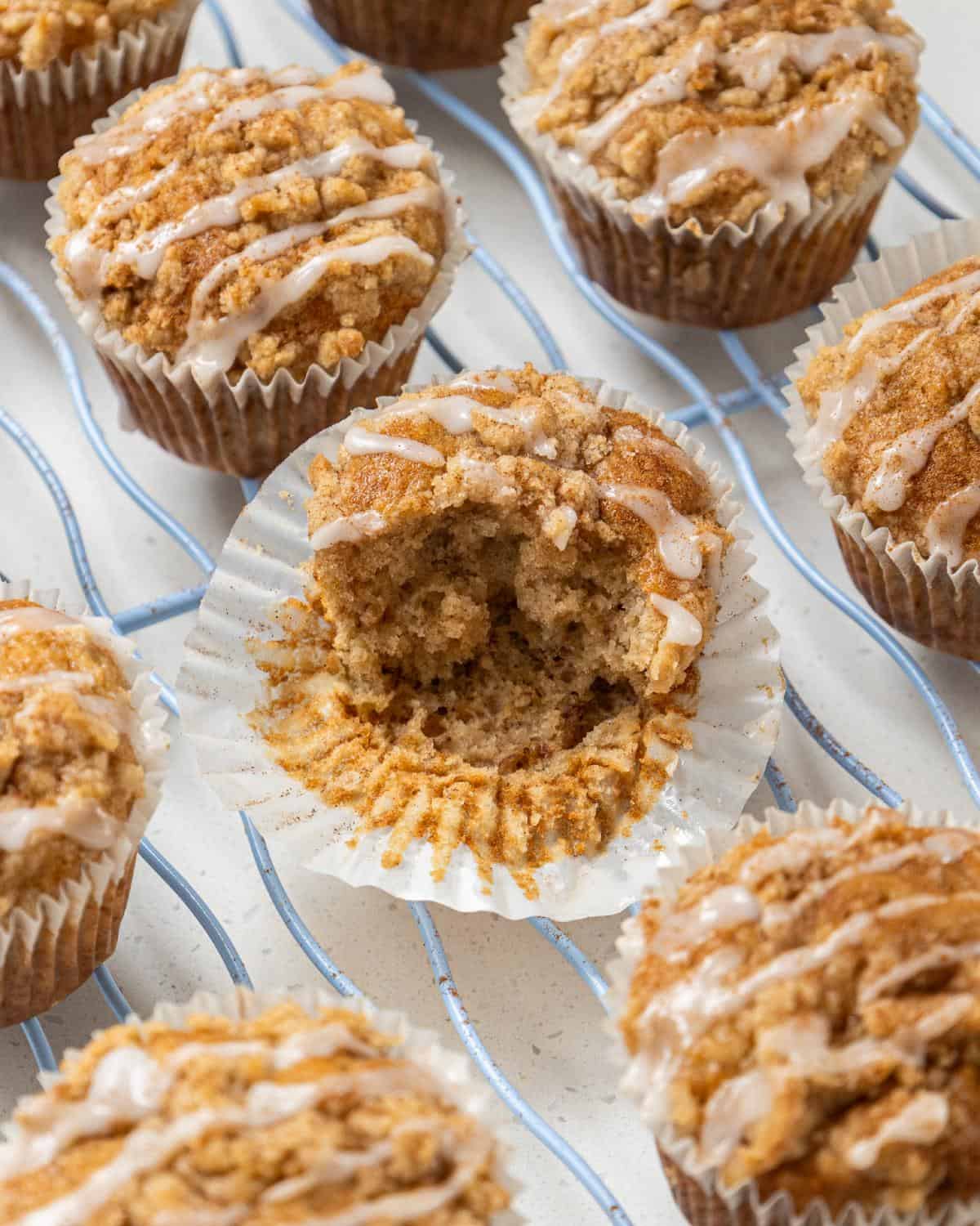 A batch of streusel-topped muffins drizzled with icing, one of which has a large bite taken out of it, sitting on a wire cooling rack.