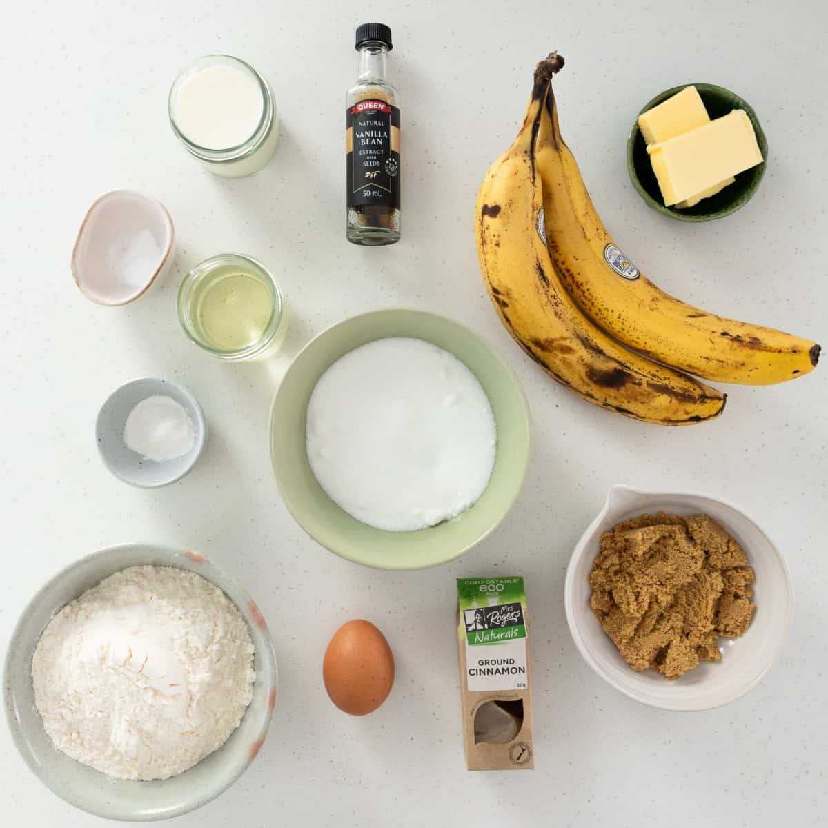 Ingredients for banana bread including flour, two bananas, white and brown sugar, butter, an egg, oil, milk, vanilla bean extract, cinnamon, salt, and baking soda arranged on a white surface.