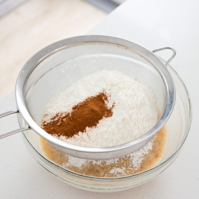 Banana Cinnamon Crumble Muffins A metal mesh sieve containing flour and cinnamon held over a glass mixing bowl with liquid ingredients.