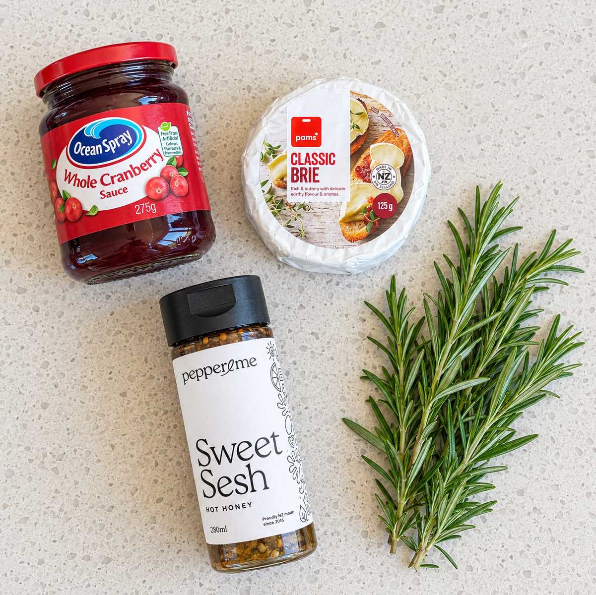 A jar of Ocean Spray whole cranberry sauce, a round of Pams classic brie, a bottle of Pepper & Me Sweet Sesh hot honey, and a sprig of fresh rosemary.