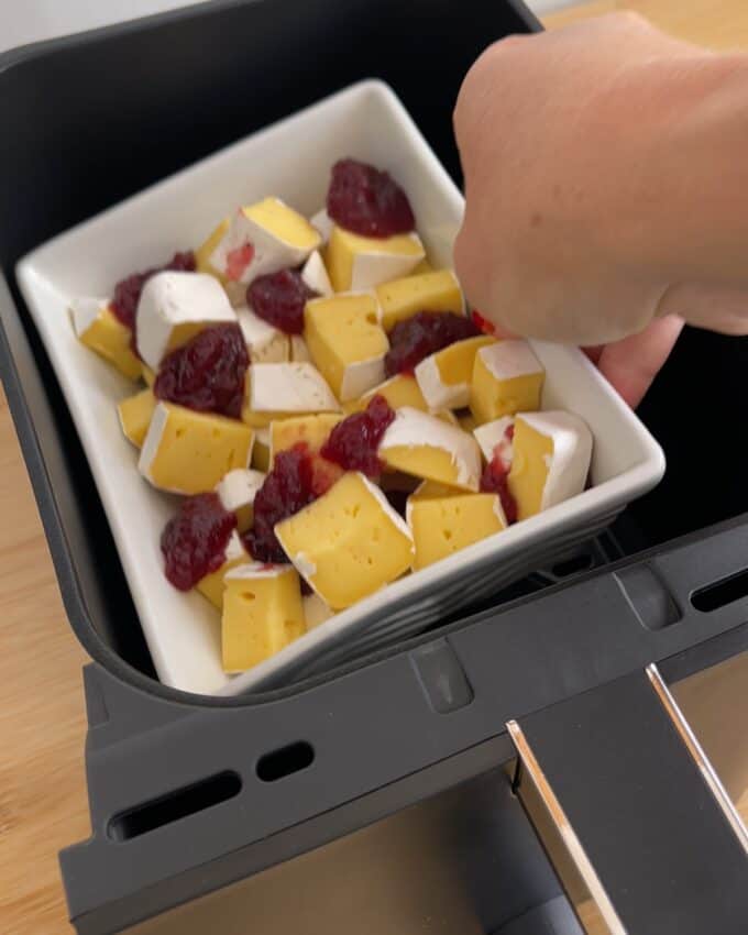 Air Fryer Cranberry Brie Dip A hand places a white square dish filled with cubed Brie topped with cranberry sauce into an air fryer basket.