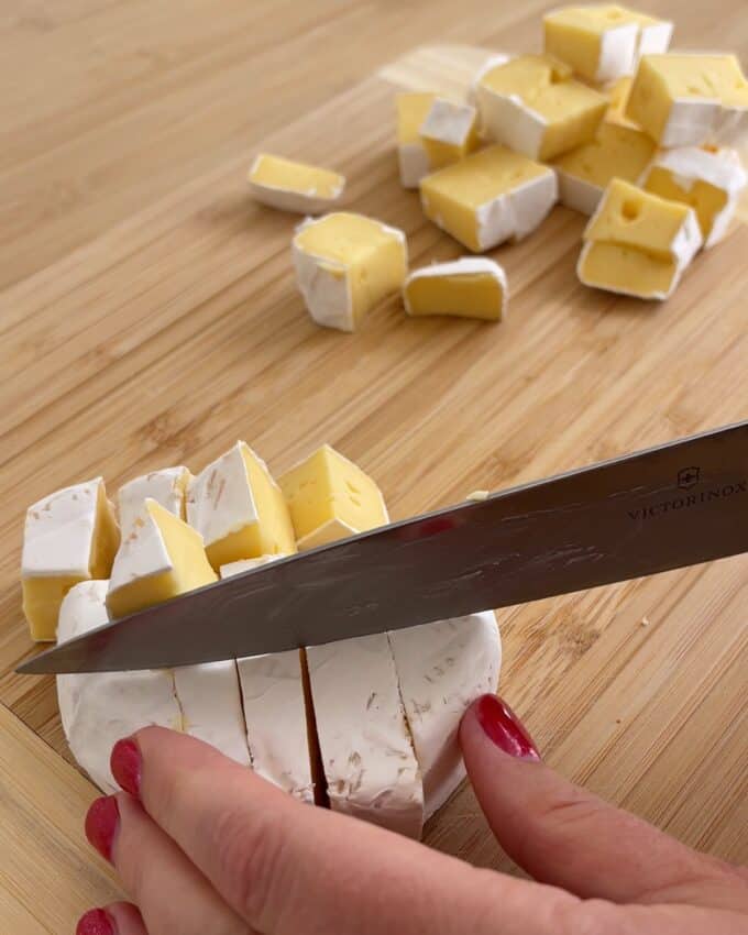 Air Fryer Cranberry Brie Dip A hand with red nail polish uses a knife to cut a wheel of brie on a wooden cutting board, with additional pre-cut pieces nearby.