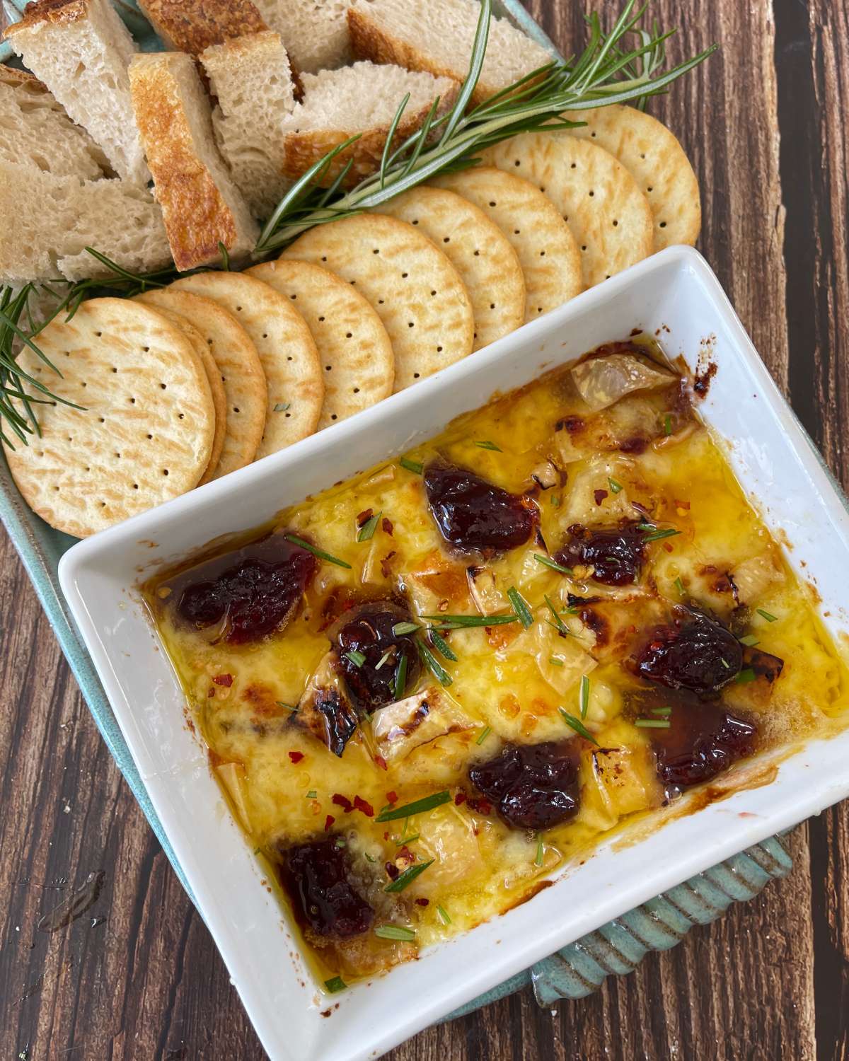 A rectangular baking dish filled with baked brie, topped with dark fruit preserves and rosemary, served with sliced bread and round crackers.
