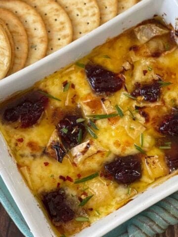 Baked brie in a rectangular dish topped with cranberry sauce and herbs, served with a side of round crackers.